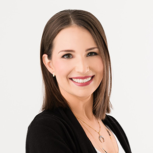 Woman with straight brown hair, wearing a black cardigan and gold necklaces, smiles at the camera against a white background.
