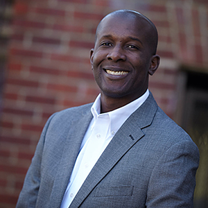 A man in a gray suit jacket and white shirt smiles while standing in front of a brick wall.