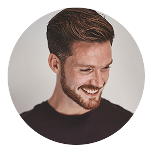 A man with short brown hair and a beard is smiling and looking down, wearing a dark shirt against a plain background.