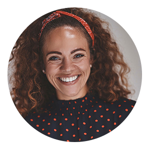 Woman with curly hair and a red headband smiling, wearing a navy blue shirt with red polka dots.